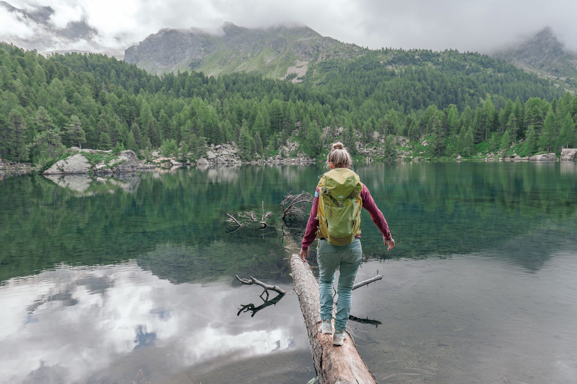 Eine Frau mit Rucksack überquert einen See auf einem Baumstamm.